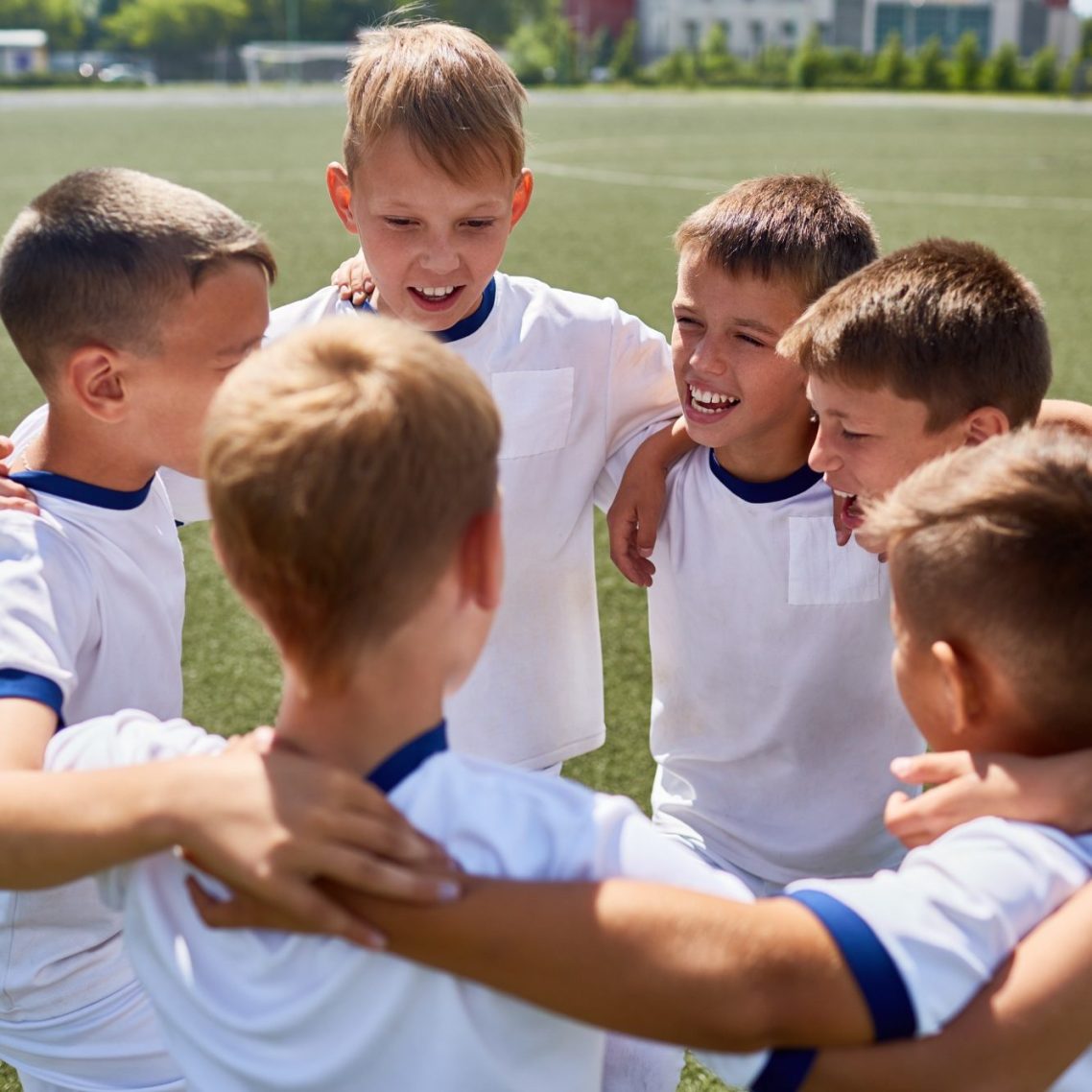Sechs Jungen in weißen Fußballtrikots umarmen sich und lächeln gemeinsam.